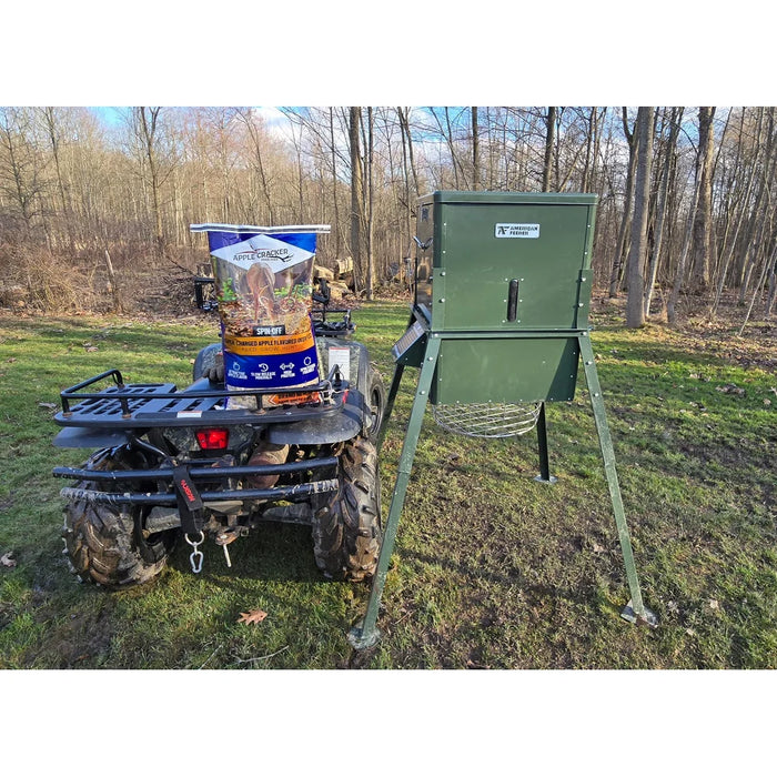 Green American Feeder AF300 Wildlife Feeder on tripod legs next to an ATV carrying a bag of deer feed in a wooded clearing