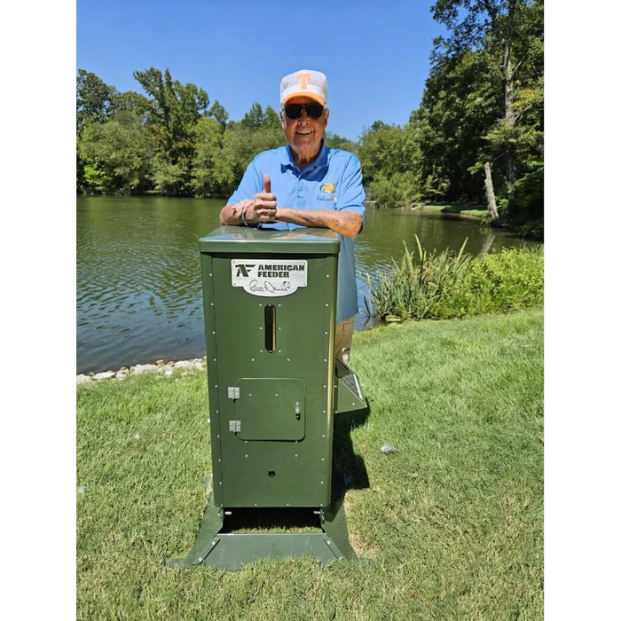 Bill Dance standing beside the American Feeder H125 Lake Feeder on a grassy bank next to a calm pond