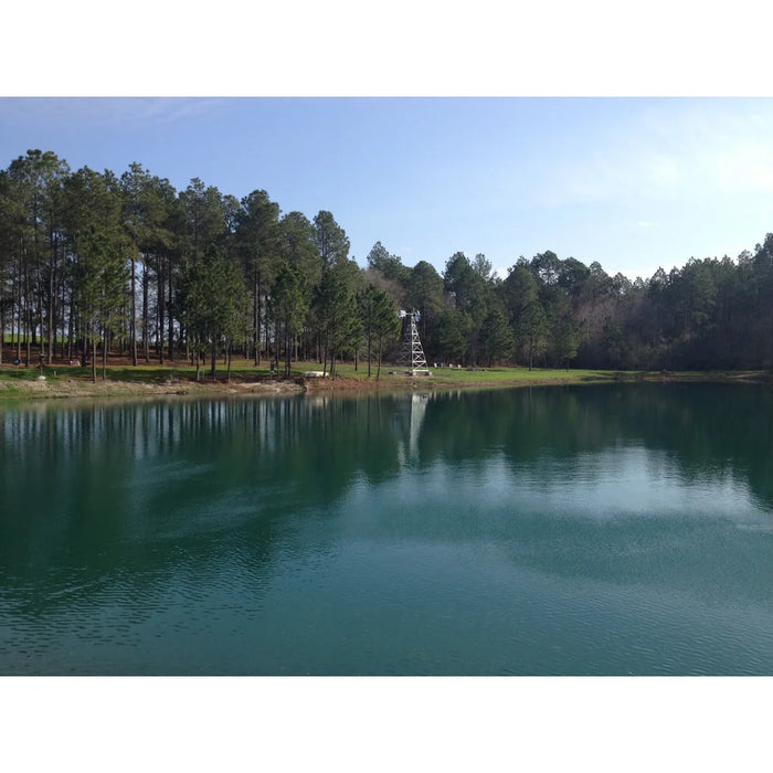 Outdoor Water Solutions Steel Tower Windmill Pond Aerator installed across a clear green pond lined with pine trees
