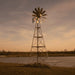 Full front view of the Outdoor Water Solutions 3-Leg Metal Windmill Pond Aerator beside a pond at dusk