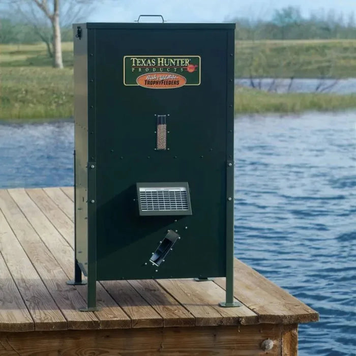 Texas Hunter Solar Fish Feeder for Pond on short legs mounted on a wooden dock beside open water with front solar panel grille