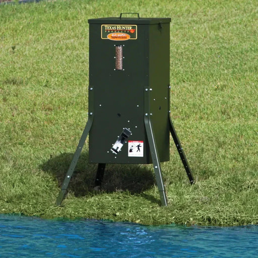 Texas Hunter Solar Pond Fish Feeder standing on splayed legs on a grassy pond bank next to blue water with feed window full of pellets