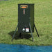 Texas Hunter Solar Pond Fish Feeder standing on splayed legs on a grassy pond bank next to blue water with feed window full of pellets
