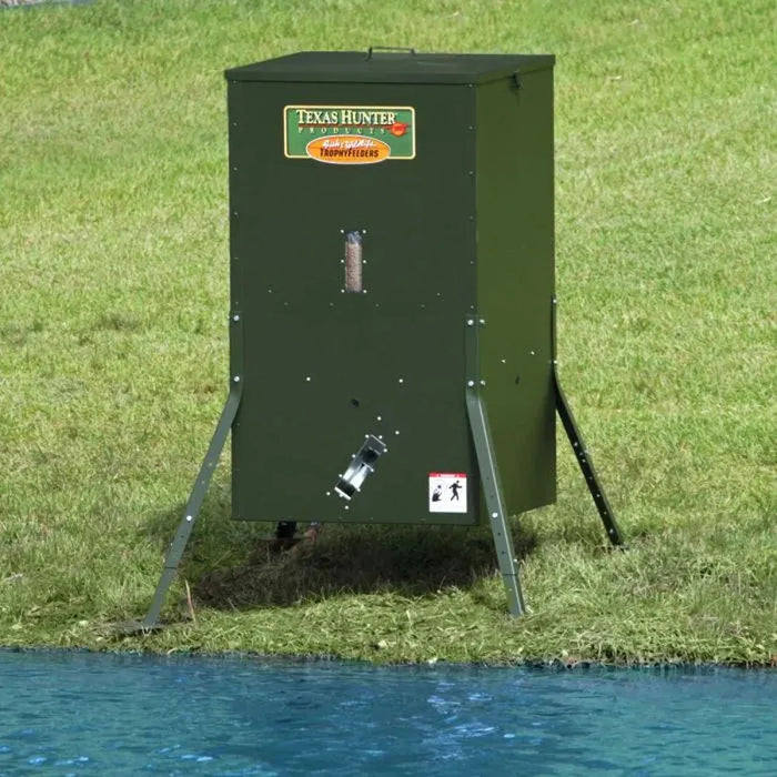 Texas Hunter Automatic Solar Fish Feeder for Pond on stationary legs beside blue pond water on a grassy bank, angled view