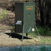 Texas Hunter Solar Fish Feeder for Pond on tall legs at a wooded pond edge with side solar panel and feed scattered below the chute